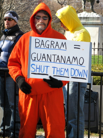 Man in orange jumpsuit holds sign: 'Bagram = Guantanamo/Shut Them Down'