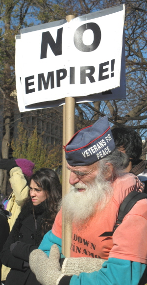 Old man, wearing a Veterans for Peace cap, holds sign: 'No Empire!'